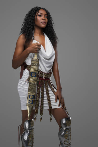 Stunning afro-American beauty with curly hair and adorned in a white Greek tunic, embellished with ornate belt and leg greave, poses with a gladius on a grey background