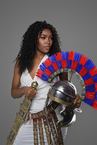 Beautiful dark-skinned model dons a white Roman tunic with a decorated belt and greaves, holding a gladius and Roman centurion helmet on a neutral grey background