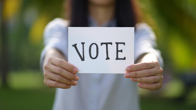 An Asian woman stands outdoors in a public park, holding a white paper card with the handwritten word 'VOTE'