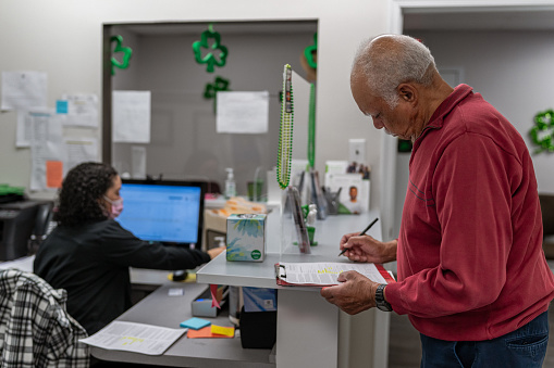 A senior man stands at a dental office reception desk and completes paperwork before his appointment. The medical clinic is decorated with green clovers in celebration of St. Patrick's Day.