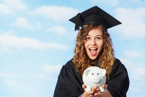 Beautiful young woman wearing mortar board and academic gown gasps in amazement as she holds a cute piggy bank.