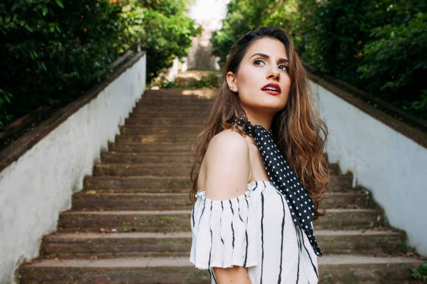 Young beautiful brunette woman standing at stairs looking away. Wears a in white off-the-shoulder dress and navy blue necklace. Summer, vacations concept.