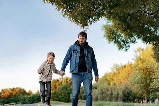 Caucasian bearded man with his little son walking in park holding hands on autumn day. Image with selective focus