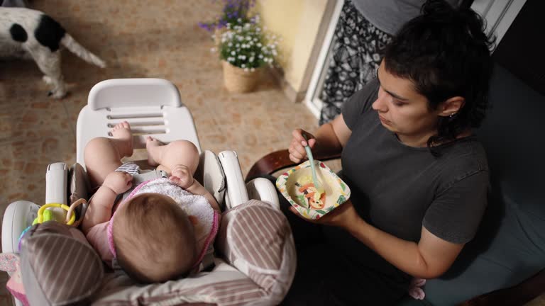 On the balcony, young mother feeding her adorable baby girl with baby food, while she seating in the car seat