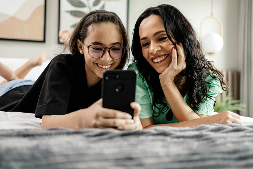 Mother and teenage daughter using smartphone together lying in bed at home. Latina mother and daughter looking at phone watching social media videos while relaxing in bedroom. family and technology