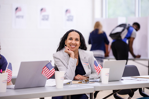 The mature adult polling place volunteer rests her head on her hand and smiles for the camera during a break.
