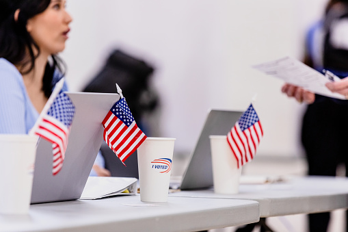 The focus of the photo is on the American flags at the volunteer table.  In the background, the polling place volunteer answers questions about the ballot.
