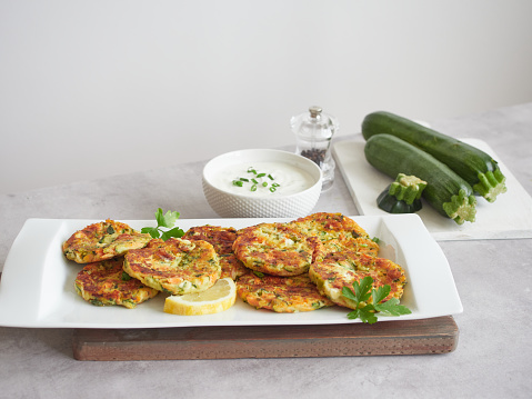 Crispy and golden zucchini vegetable pancakes served on a platter on the table next to a bowl of yogurt cream, a pepper shaker and raw green zucchini on a cutting board. Healthy vegetarian meal.