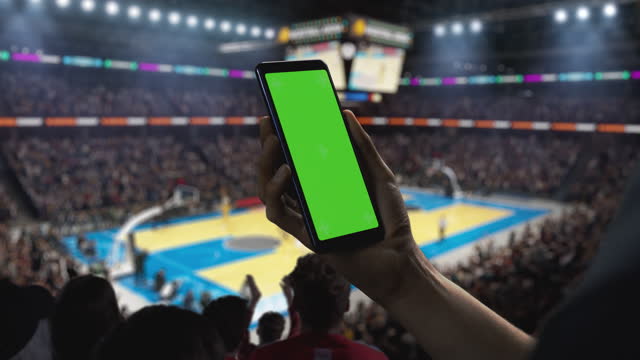 Basketball Championship: Person's Hand Holding a Smartphone with a Green Screen Display. Sports Fans in an Arena Cheering for a Team to Win. Template to Use for Social Media, Scores, Results, Betting