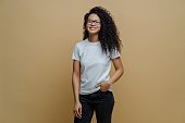 Positive ethnic student, free after passing exam, broad smile, hand in jeans pocket, transparent glasses. Brown background. People, lifestyle concept.