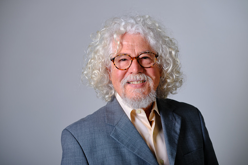 front view portrait of a caucasian old man with white wavy hair and eyeglasses in a gray background studio