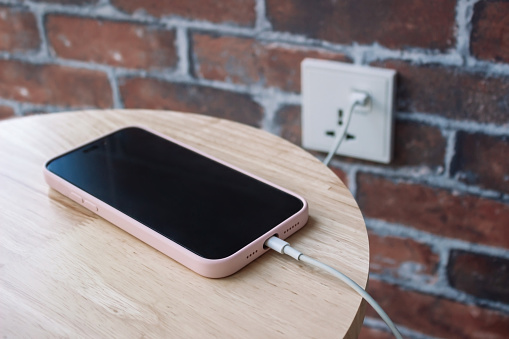 A mobile phone in a pink case charging battery by wire. A charging smartphone with cable on a wooden bedside table on a red brick wall backround