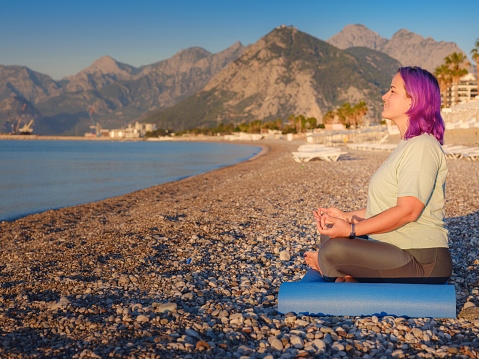 Woman Spending time outside in summer morning near the sea, meditates enjoying sound of ocean waves, sitting on seashore. Body positive , losing weight, slimming, stretching, relax and zen concept