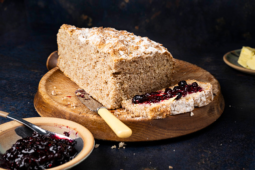 A loaf of wheaten bread on a bread board. One slice of the bread has been spread with butter and summer berry fruit jam.