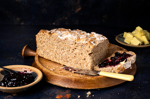 A loaf of wheaten bread on a bread board. One slice of the bread has been spread with butter and summer berry fruit jam.