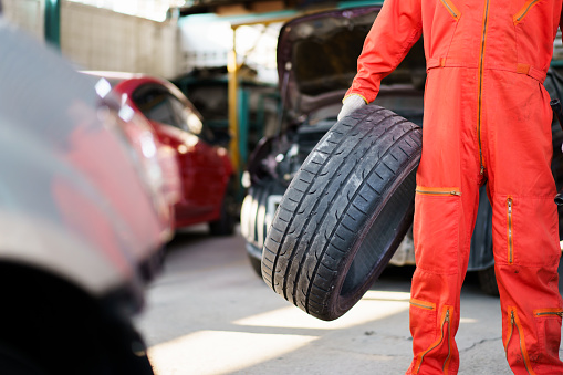 Portrait of professional Asian male vehicle technician or repairman in orange suit holding - carrying a damaged vehicle tyre to be a fixed, a repair man fixing and recap a broken tyre in garage.