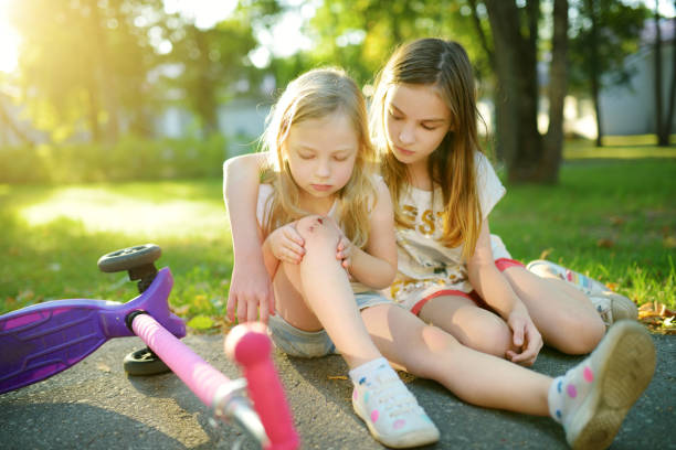 Adorable girl comforting her little sister after she fell off her scooter at summer park. Child getting hurt while riding a kick scooter. stock photo