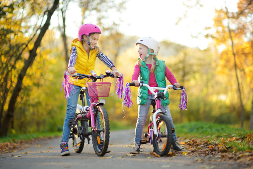 Cute little sisters riding bikes in a city park on sunny autumn day. Active family leisure with kids. Children wearing safety helmet while riding a bicycle.