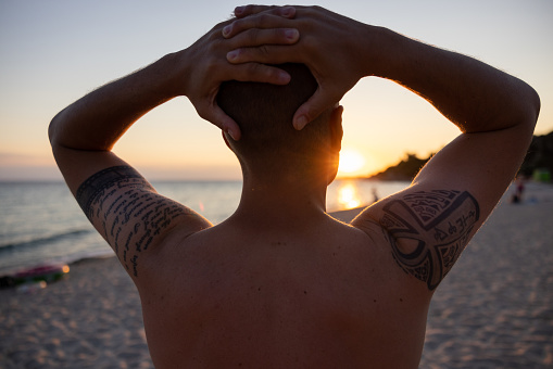 Rear-view of a young Caucasian man, admiring the sunset, on the beach, while holding a hand behind head