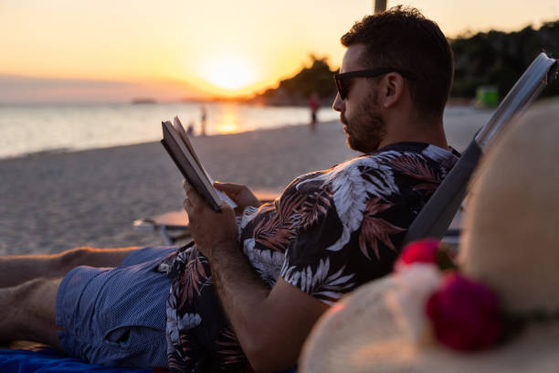 istock Young man, sitting on the beach chair, during sunset, and reading a book 1502960500