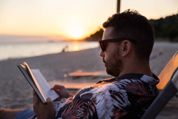 istock Young man, sitting on the beach chair, during sunset, and reading a book 1502960520