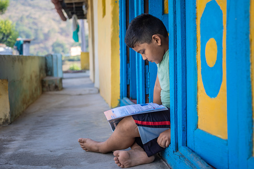 Elementary age child reading his school book. He is sitting at his village house door. Child education concept.