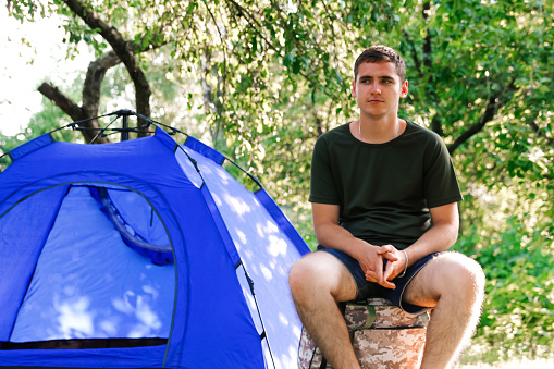Man tent summer. Defocus young man tourist sitting in touristic tent at the beach. Camping, tourism and travel concept. Green forest. Out of focus.