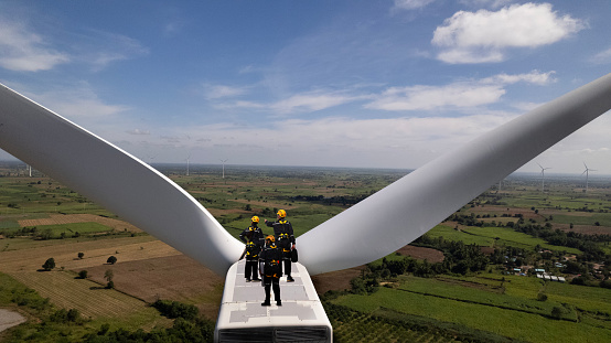 Wind Turbine Maintenance team working on top of wind turbine stands. group of technical engineers wearing safety suits and hardhat checking the wind turbine motor on the top of the power supply.