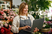 Small flower shop owner florist wearing apron working on laptop and taking online orders in store