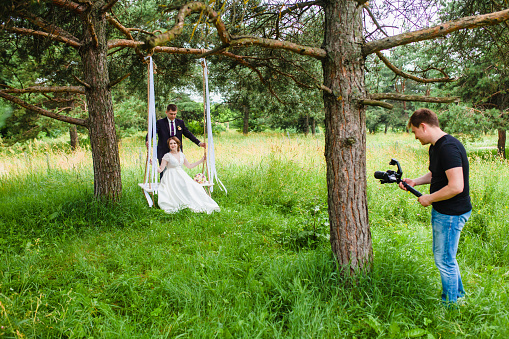 Professional wedding videographer shoots a film with the newlyweds on a rope swing in a pine forest