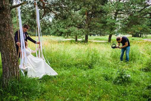Professional wedding videographer shoots a film with the newlyweds on a rope swing in a pine forest
