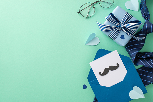 Stylishly modern Father's Day concept. Overhead shot of postcard with mustaches, gift box, necktie, spectacles, and accessories on a teal background with an empty space for text