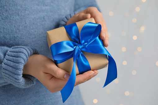 Woman holding gift box with blue bow against blurred festive lights, closeup and space for text. Bokeh effect