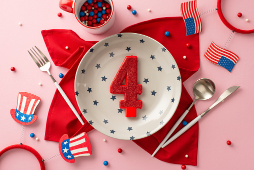 Fourth of July festivities menu. Top-view perspective of table setup, dinner plate, utensils, cup, napkin, number 4 cake candle, party headbands, American flags, sprinkles, on light pink background