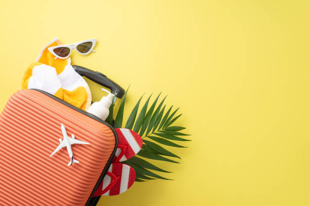 istock Get ready for summer! Top view of suitcase, small plane model, beach accessories, sunglasses, sunscreen, flip-flops, towel, and palm leaf against yellow background. Advertise your vacation deals here 1503018753