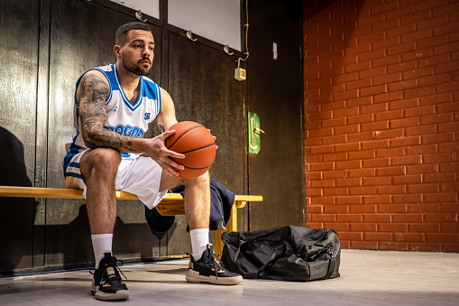 Male basketball player sitting in locker room and holding ball while waiting for sport match front side view wide shot