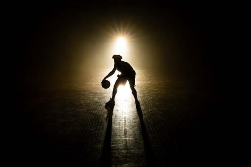 Male basketball player silhouette dribbling ball at dark court illuminated with lights wide shot