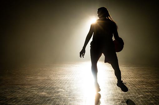 Female basketball player silhouette training at dark indoor court illuminated with lights wide shot