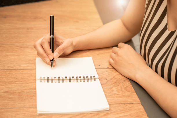 istock Woman hand is writing on a blank notepad with a pen on wood background. 1503208634