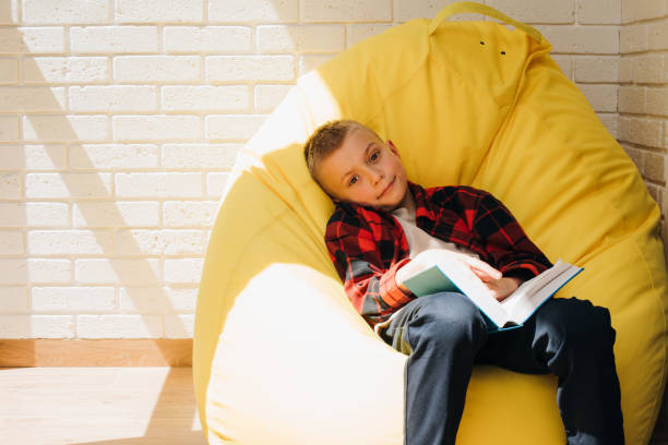 A school-age boy sits on a yellow bean bag with a book in his hands. Children reading stock photo