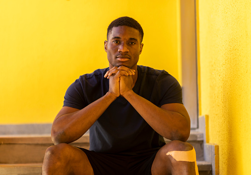 Portrait African American Young Sportive Man Sitting on Stairs Holding Hands Below Chin, With Elastic Knee Bandage, Looking At Camera On Yellow Background Outdoor. Horizontal Plane. High quality photo
