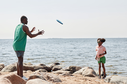 African American dad throwing plate to his little daughter, they playing outdoor game on the beach