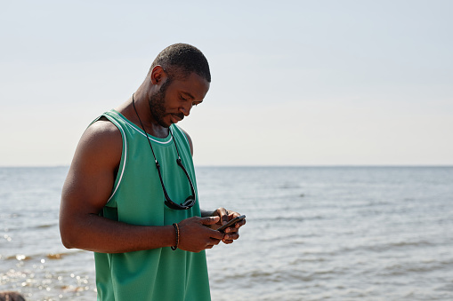 African American young man texting message on smartphone standing on the beach