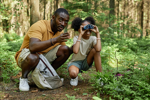 African American dad teaching his daughter to hiking, she looking through the binoculars during their journey