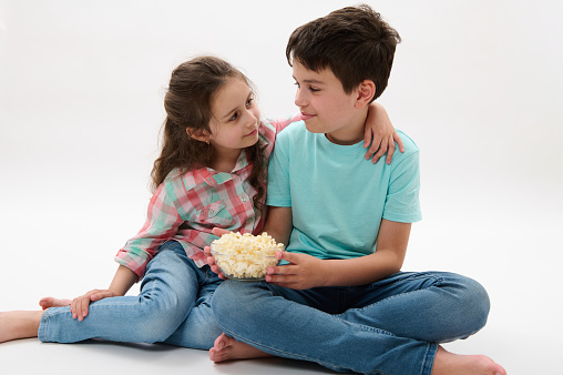 Adorable nice kid girl in plaid shirt and casual denim, gently hugging her older brother serving her a bowl of delicious popcorn while film screening, isolated on white studio background. Happy family
