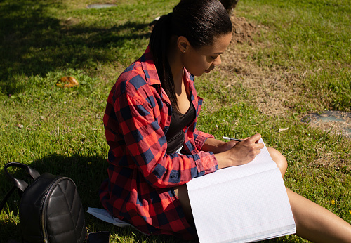 College student girl writing in notebook outdoors, studying and preparing to exam while spending free time in park.
