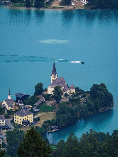 Aerial view of the church of Maria Worth (Maria Wörth) at Worthersee (Wörthersee) with boat Aerial view of the church of Maria Worth (Maria Wörth) at Worthersee (Wörthersee) in the summer with a boat as seen from the Pyramidenkogel lookout tower maria woerth stock pictures, royalty-free photos & images