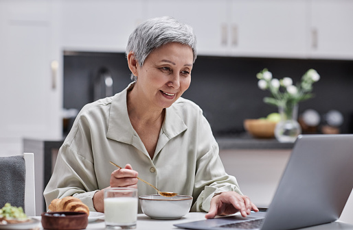 Portrait of smiling senior woman using laptop and working while enjoying breakfast at home, copy space