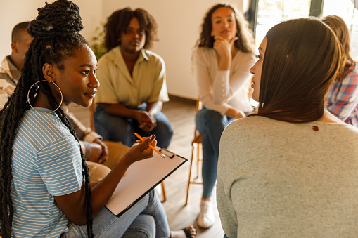 Selective focus shot of female therapist sitting in circle, holding a clipboard to take notes and talking during a group therapy session.