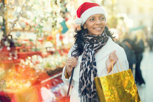 Hispanic woman walking at christmas fair with gift bag in hand and looking in camera.
